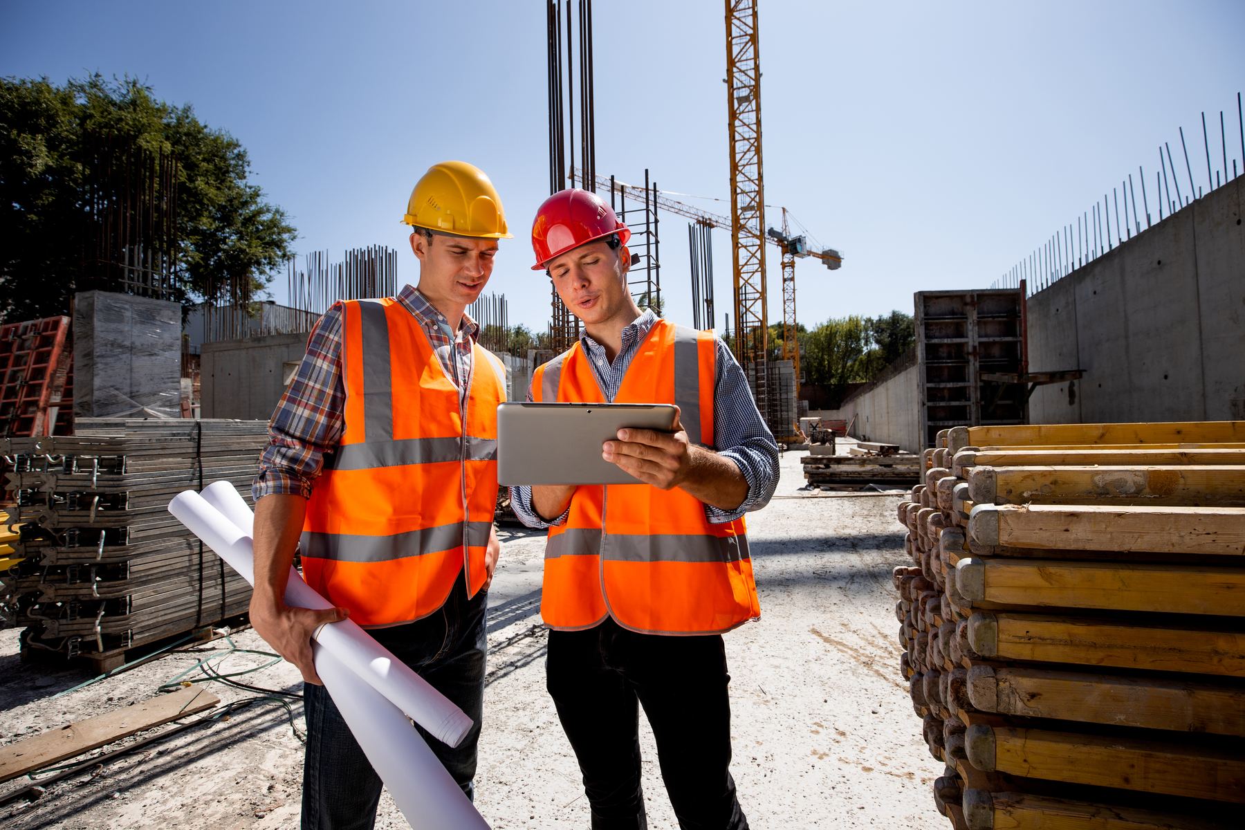 Zwei Handwerker auf der Baustelle prüfen eine digitale Checkliste auf dem Tablet, eine Bauplanrolle noch in der Hand